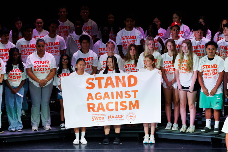 Students from across Worcester Public Schools stand on stage holding a sign that says "Stand Against Racism."