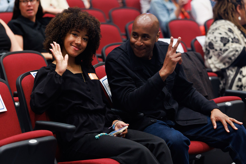A student waves at the camera while sitting in an auditorium. A man beside her gives the peace signs.