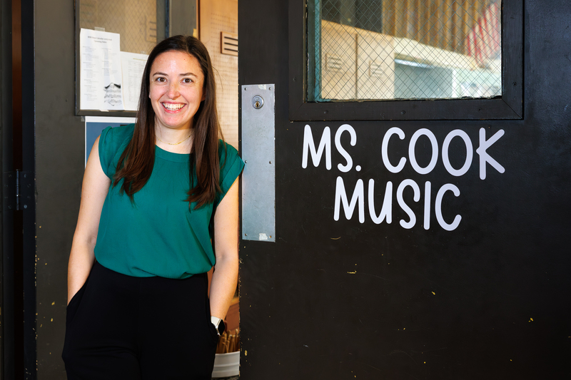 Andrea Cook stands outside of her classroom, the words 'Ms. Cook' and 'Music' are visible on the door.