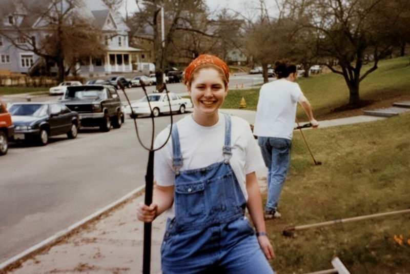 A young woman in denim overalls and a red bandana smiles while holding a pitchfork.