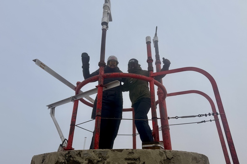 Two people in winter gear stand on a platform near antennas, with a foggy sky in the background.