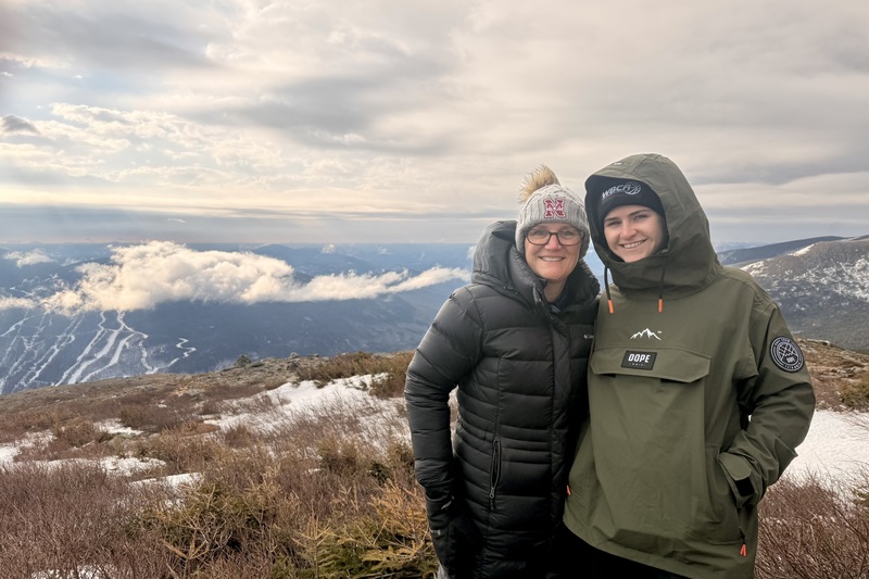Two smiling individuals in winter jackets and hats stand on a snowy mountain overlooking a cloudy sky and distant valleys.
