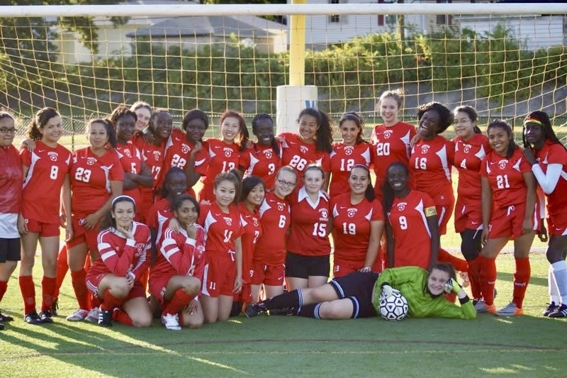 A diverse group of young women in red soccer uniforms pose for a team photo on a green field.