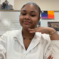 A young woman with her hand on her chin smiles in front of a whiteboard with artwork.