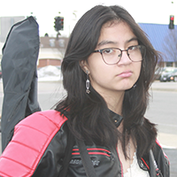 A young person wearing a black and red jacket and glasses stands outdoors with a guitar case.