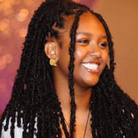 A young Black woman with long, dark locs smiles warmly, wearing gold earrings.