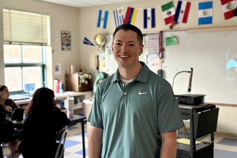 A man in a teal polo shirt gestures while speaking to students in a classroom.