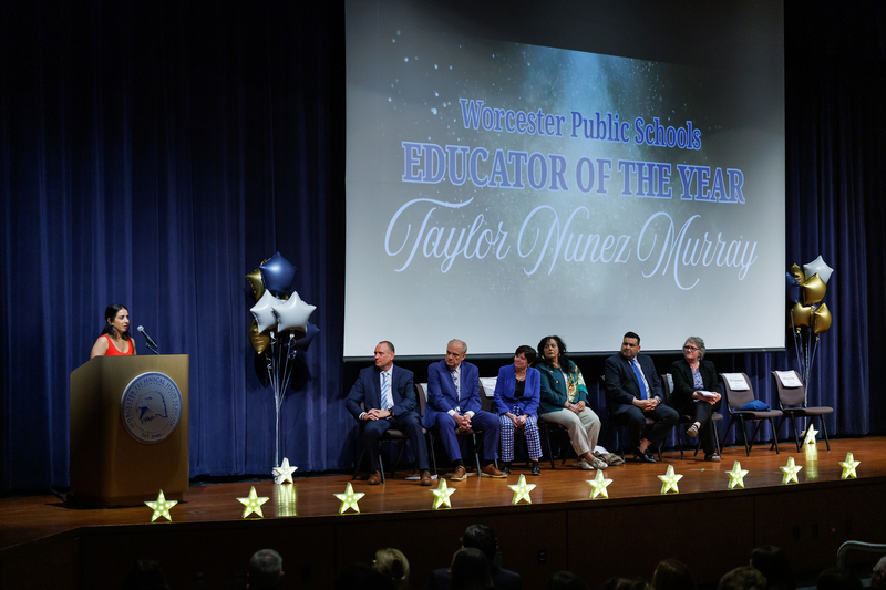 Taylor Nunez gives a speech while elected officials look on. The projection says "Worcester Public Schools Educator of the year"
