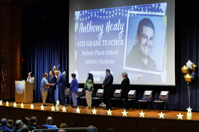 An adult shakes the hand of an elected official while walking across stage, a projection of their head shot is visible.