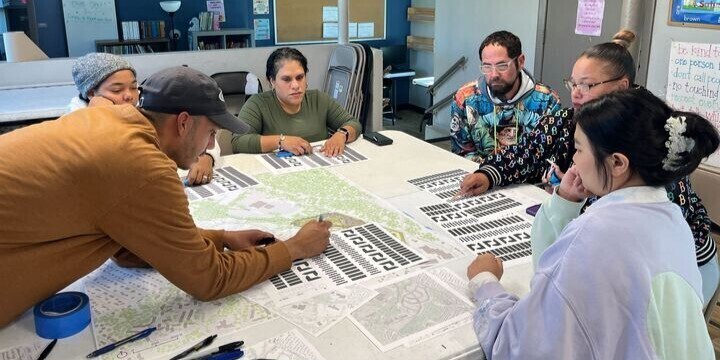 A diverse group of people collaborate around a table, examining maps and documents.