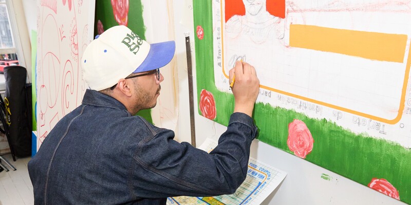 An artist in a denim jacket and baseball cap sketches on a large canvas with floral elements.
