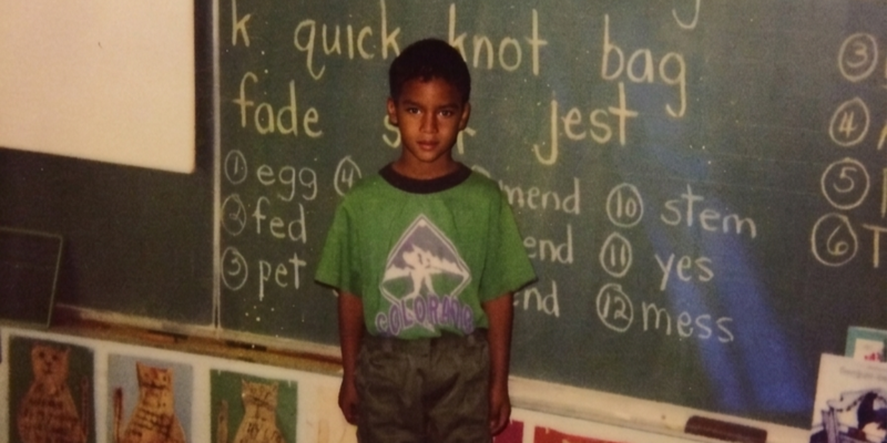 A young boy stands in front of a chalkboard with words like 'quick', 'knot', and 'bag' written on it.