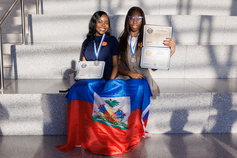Two students sit on a stairwell with a flag drapped over their legs while holding their certificates following a celebration ceremony.