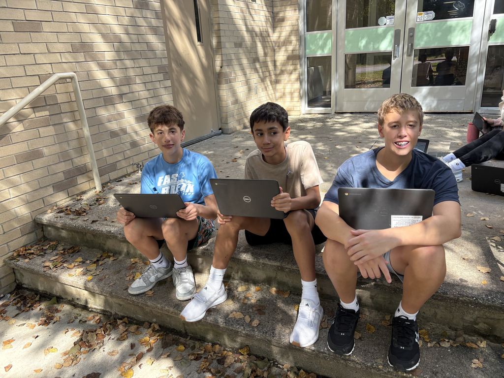 3 students sit on the steps of Eisenhower Middle School with their chromebooks and smile at the camera. 