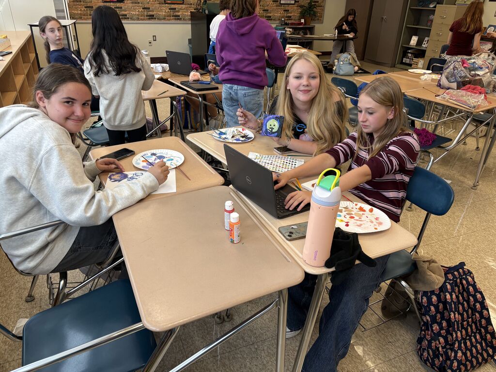 2 students smile for the camera while one searches on her chromebook for inspiration for her craft.