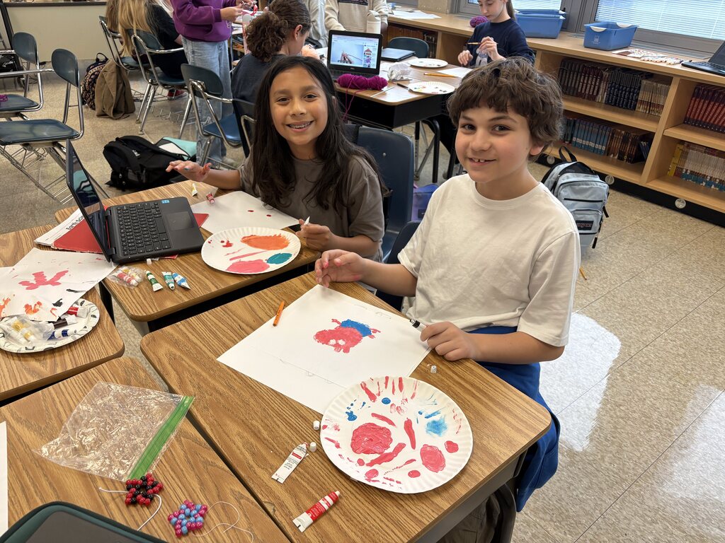Two students sit at desks with paper plates of paint in front of them.