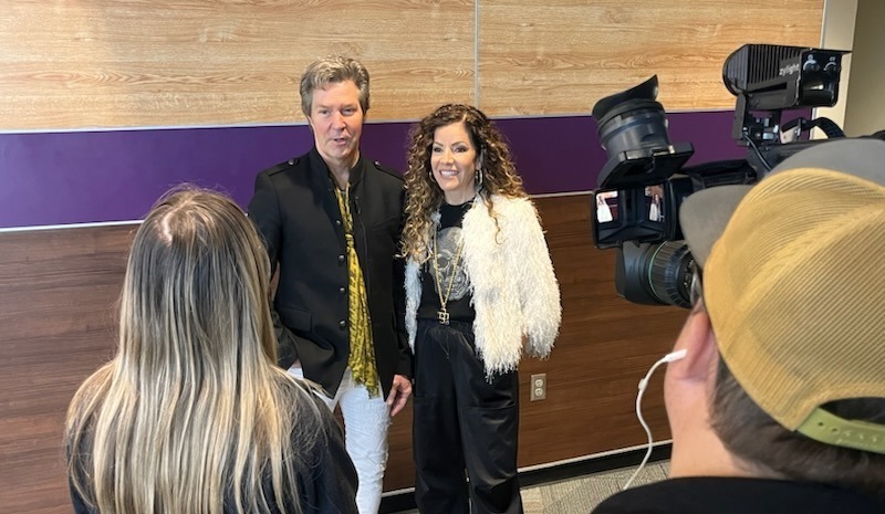 "A man and a woman, both dressed stylishly, stand in front of a wooden and purple wall while being interviewed by a small group. The man is wearing a black jacket with a patterned yellow scarf, white pants, and sneakers, while the woman wears a fluffy white jacket over a black outfit with gold accessories. Two people are seen from behind, one with long blonde hair and another wearing a yellow baseball cap, both facing the interviewees. A professional video camera is capturing the interview from the right side of the frame."