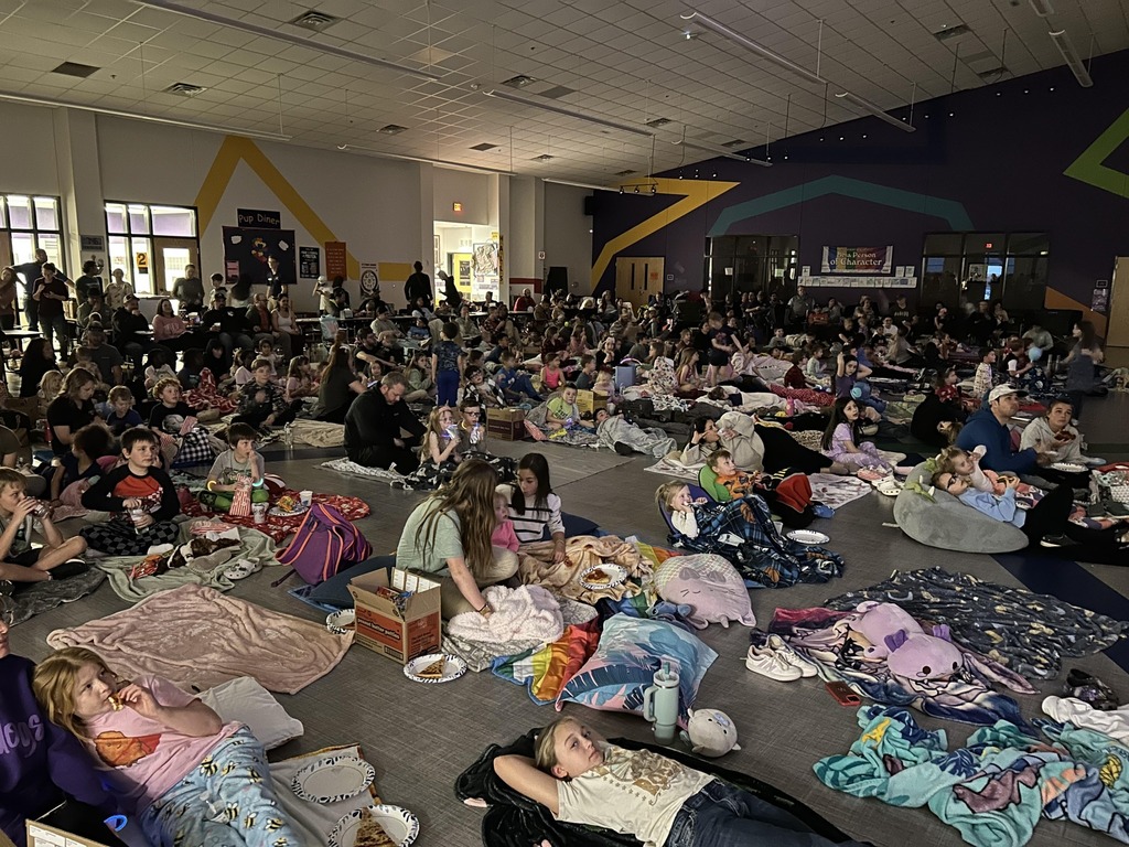 A large group of children and families gather in a dimly lit school cafeteria or multipurpose room for a movie night. Many children are sitting or lying on blankets, bean bags, and pillows, dressed in pajamas, while eating snacks like pizza and popcorn. Some children are cuddled up with stuffed animals. The background features cafeteria tables with additional attendees, colorful geometric wall designs, and a sign that reads 'Pup Diner.' The atmosphere is cozy and filled with excitement as families enjoy the event together.