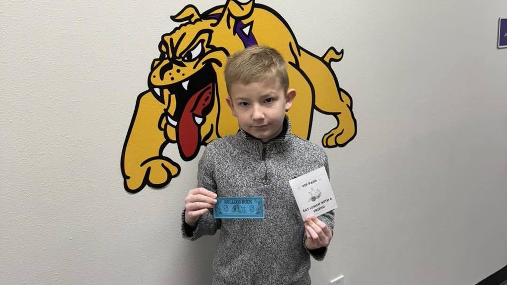 A student at Wylie Intermediate East stands in front of a bulldog mascot mural, holding a blue 'Bulldog Buck' and a white VIP pass for eating lunch with a friend.