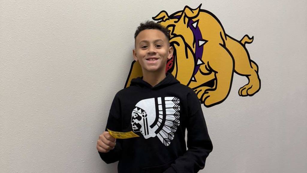 A student at Wylie Intermediate East smiles while holding a yellow reward ticket, standing in front of a bulldog mascot mural.