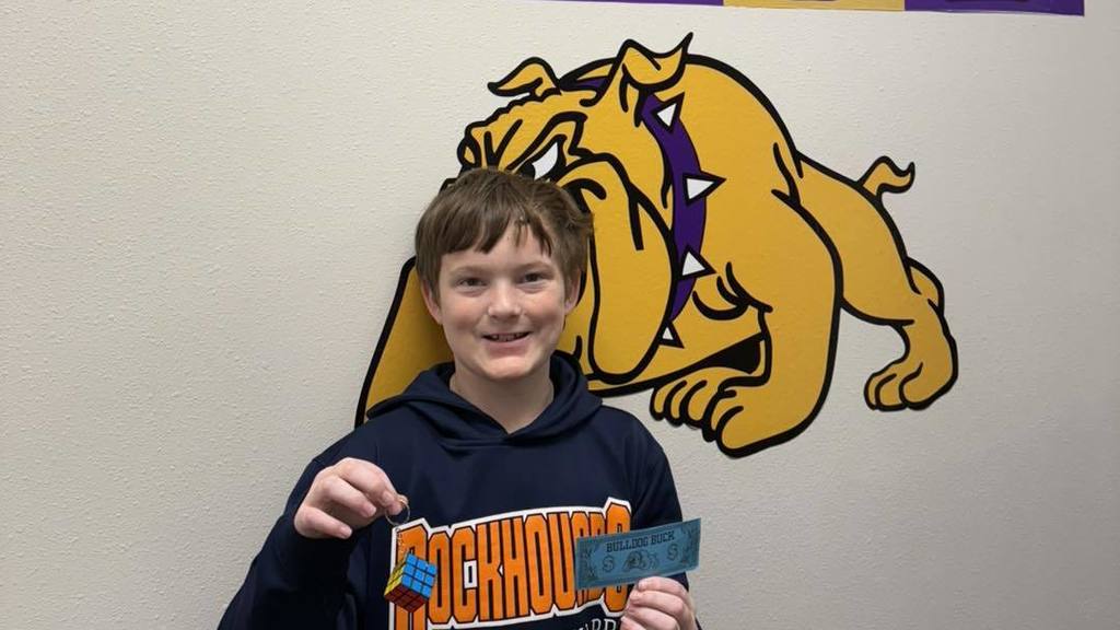 A student at Wylie Intermediate East smiles while holding a blue 'Bulldog Buck' and a small Rubik's Cube keychain, standing in front of a bulldog mascot mural.