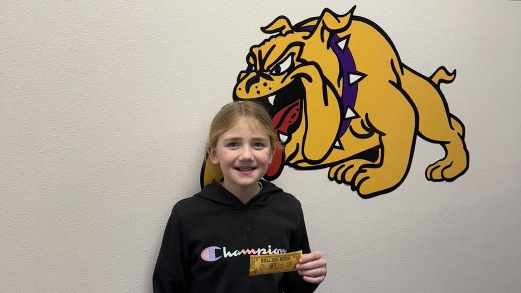 A student at Wylie Intermediate East smiles while holding a yellow 'Bulldog Buck,' standing in front of a bulldog mascot mural.