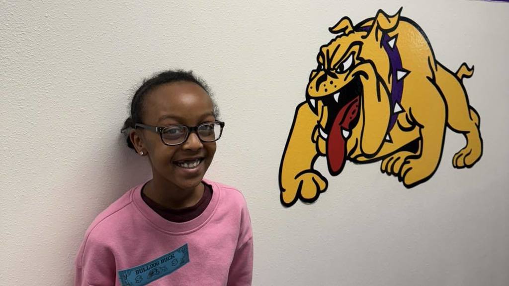 "A student at Wylie Intermediate East smiles while wearing a pink sweatshirt with a blue 'Bulldog Buck' attached, standing in front of a bulldog mascot mural.