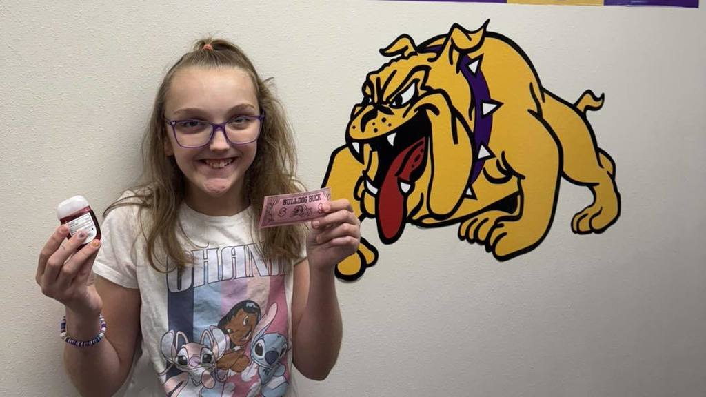 A student at Wylie Intermediate East smiles while holding a pink 'Bulldog Buck' and a small bottle, standing in front of a bulldog mascot mural