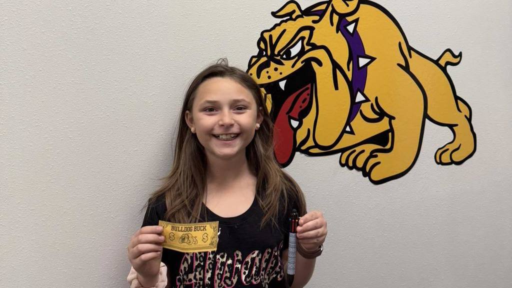 A student at Wylie Intermediate East smiles while holding a yellow 'Bulldog Buck' and a pen, standing in front of a bulldog mascot mural.