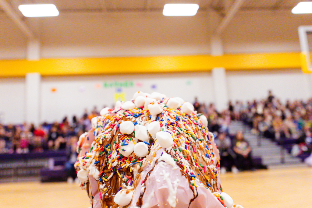 Close-up of a head covered in whipped cream, chocolate syrup, sprinkles, and mini marshmallows during a school gym event, with a blurred crowd in the background.