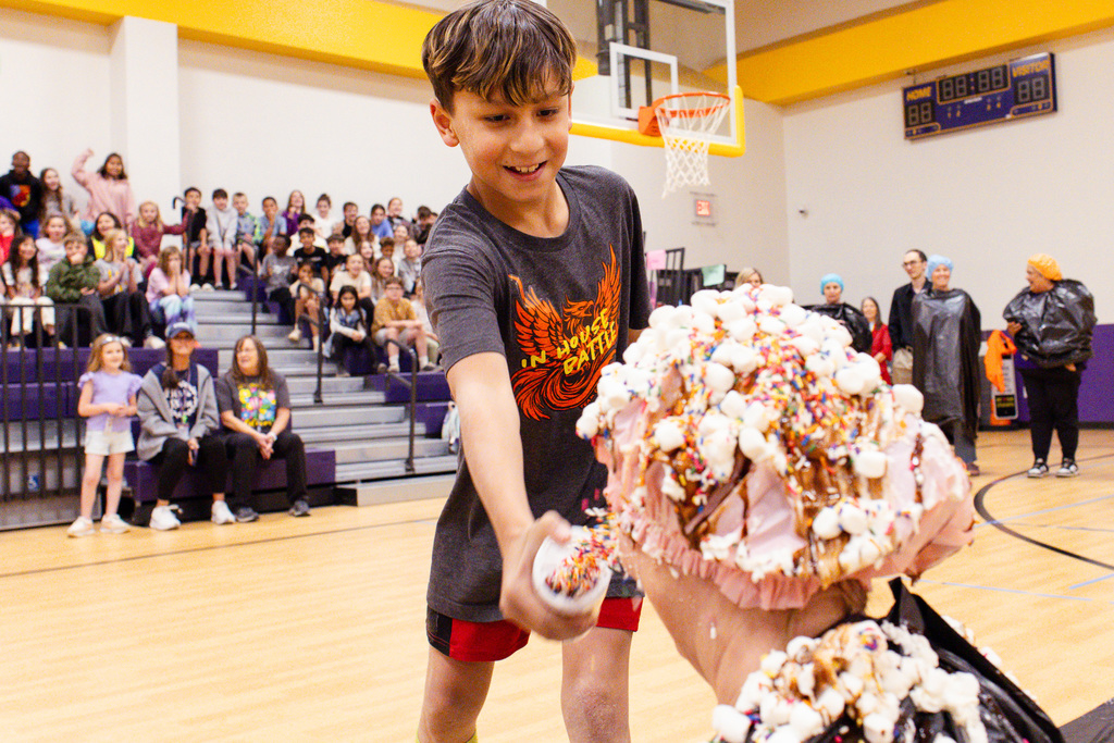 Smiling student adds whipped cream and toppings to a staff member’s head, already covered in marshmallows and sprinkles, during a school gym celebration with cheering students in the background.
