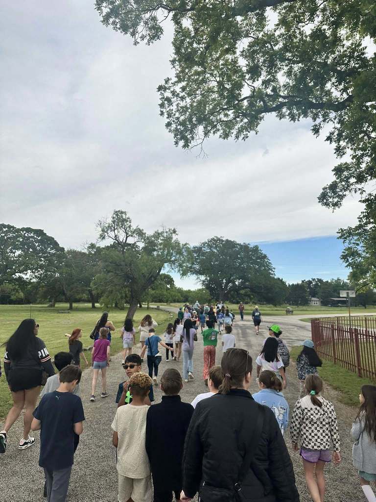 A large group of students and teachers walk along a paved path under shady trees during a field trip.