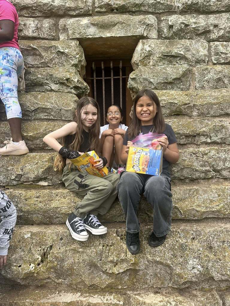 Three smiling girls hold snacks while sitting on the steps of a historic stone structure with a barred window behind them.