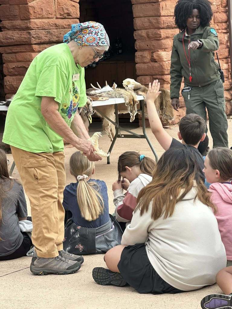 A park ranger and volunteer show animal bones and pelts to seated students, some of whom raise their hands to ask questions.