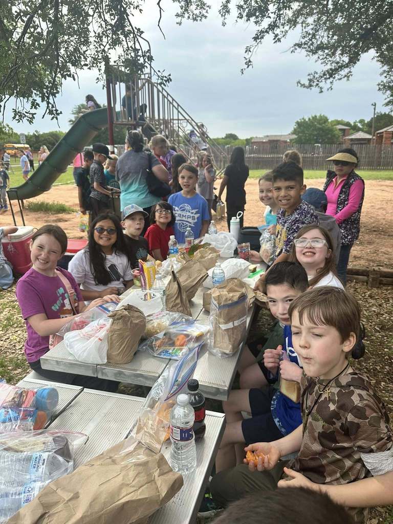 Students eat packed lunches at picnic tables in a park near a playground, surrounded by classmates and teachers.