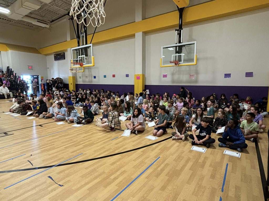 Students seated on a gym floor during a school assembly or awards ceremony.