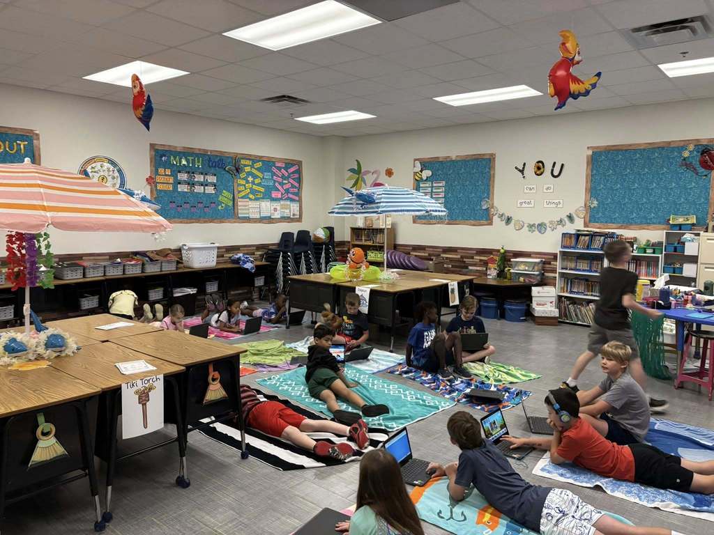 Students work on laptops while lounging on beach towels under umbrellas during a classroom beach-themed learning day.