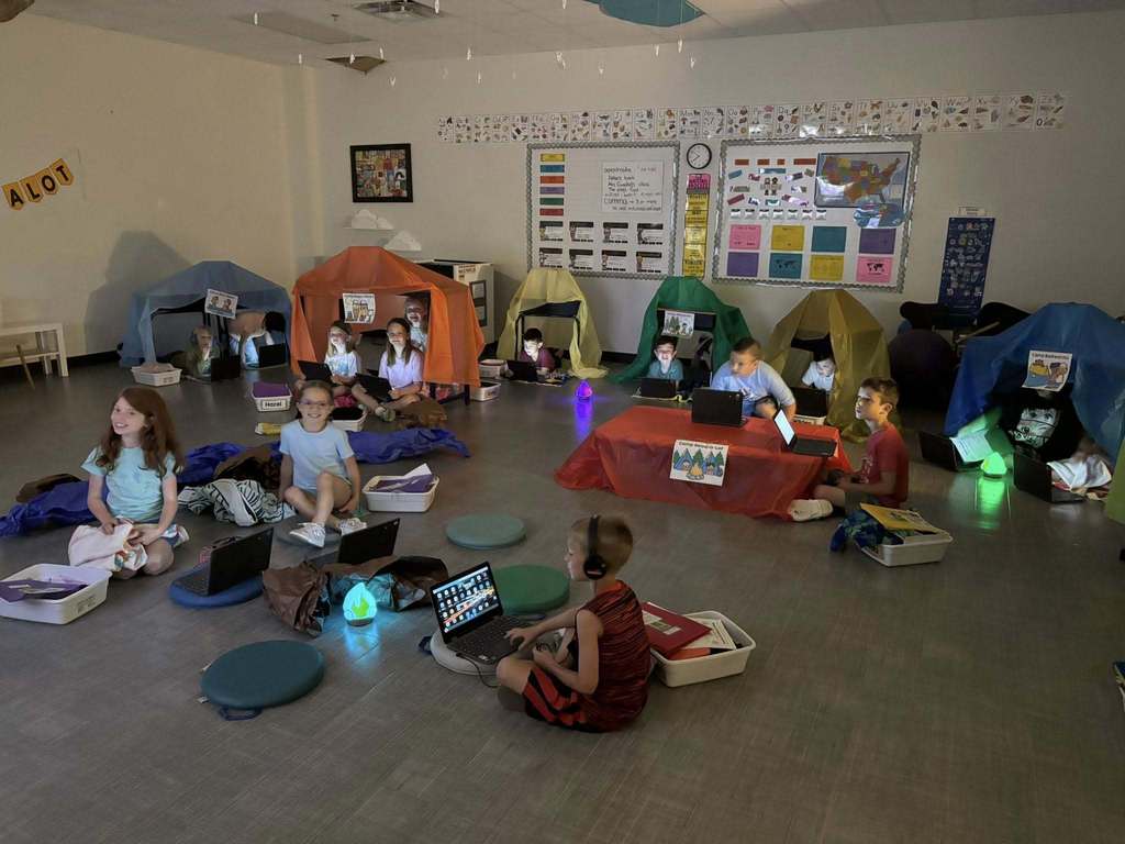 Students use laptops inside mini tents and around faux campfires during a classroom camping-themed learning activity.