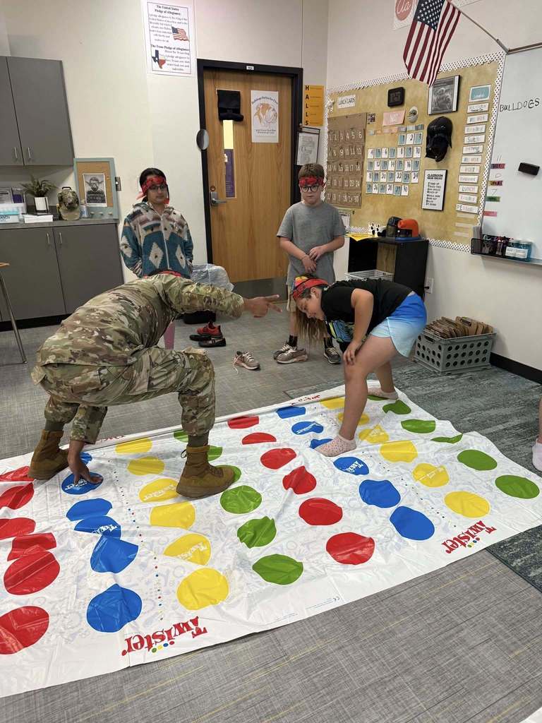 Students play a game of Twister while a service member leads the activity and classmates look on.