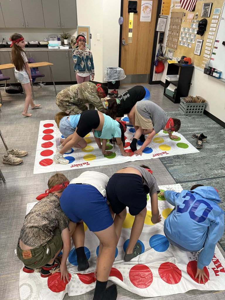 Multiple students play Twister in a classroom as a service member gives instructions near a screen showing baby photos.