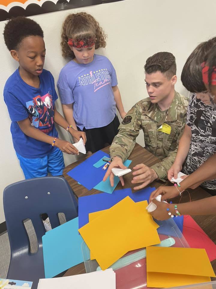 A service member sits at a table with students creating paper crafts from colorful sheets.
