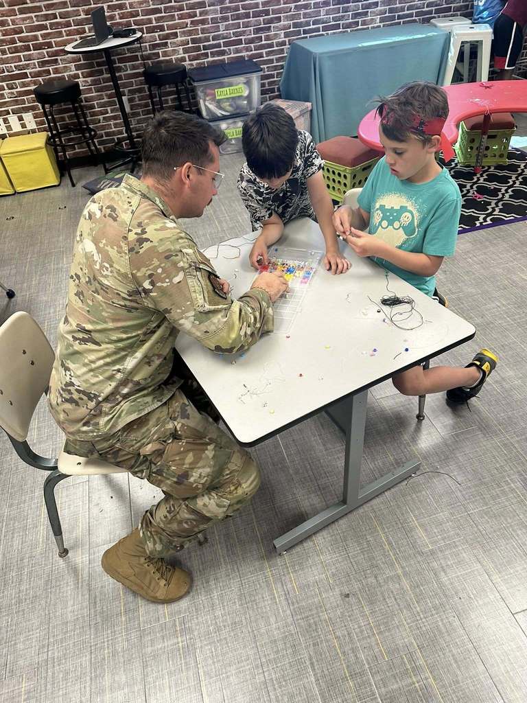 A group of students gathers around a table with a service member, working on bracelet-making.