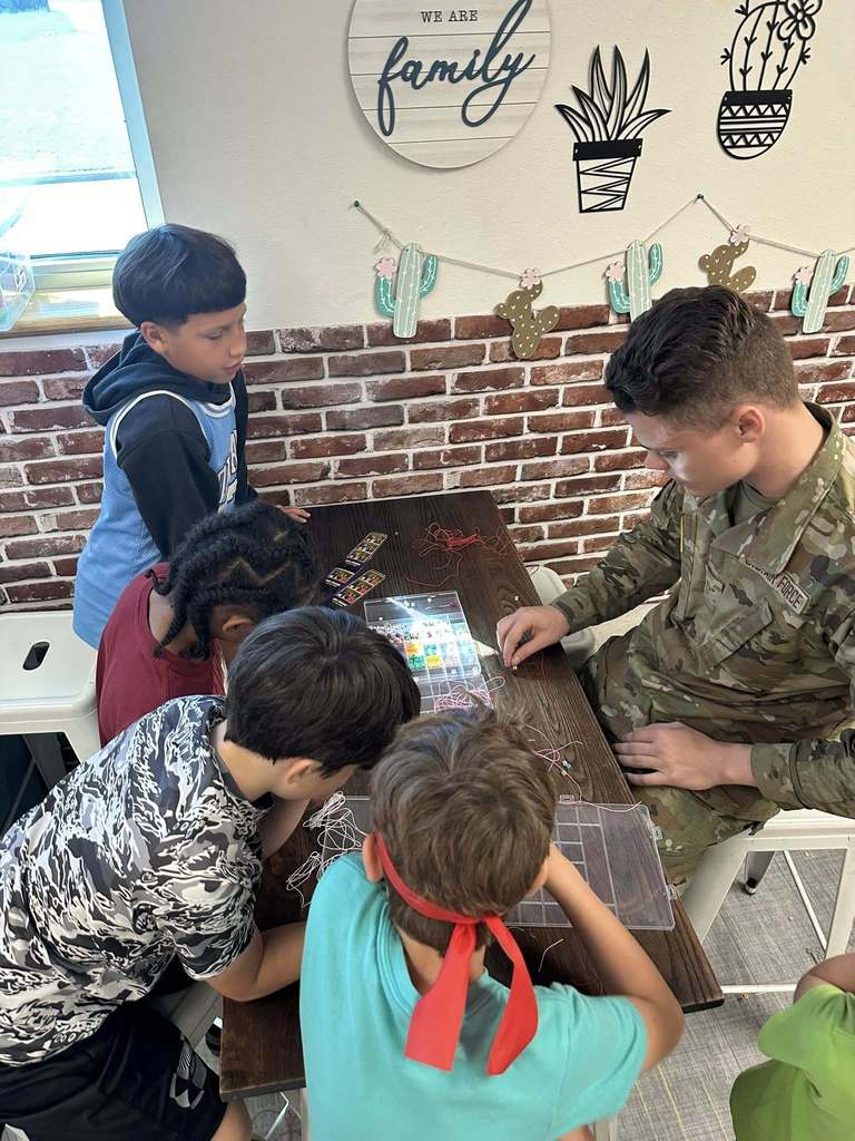 A service member helps two students with a beading craft at a classroom table.