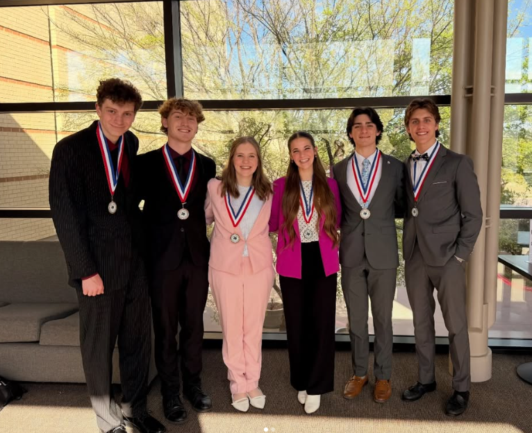 Six high school students dressed in formal attire stand side by side indoors, each wearing a medal on a red, white, and blue ribbon. They are smiling and posing in front of large windows with trees visible outside.
