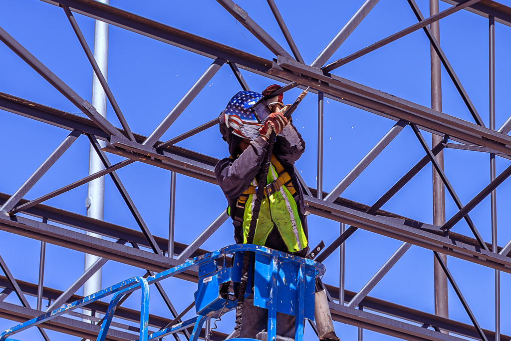 Construction worker wearing safety gear welds steel beams from a lift, assembling the structural framework of the new band expansion against a clear blue sky.