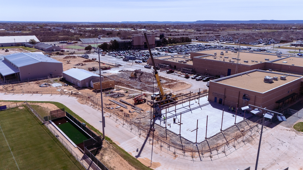 Aerial view of construction at Wylie High School showing the new band expansion in progress, with steel beams being installed by a crane on a freshly poured concrete foundation near existing school buildings and athletic fields.