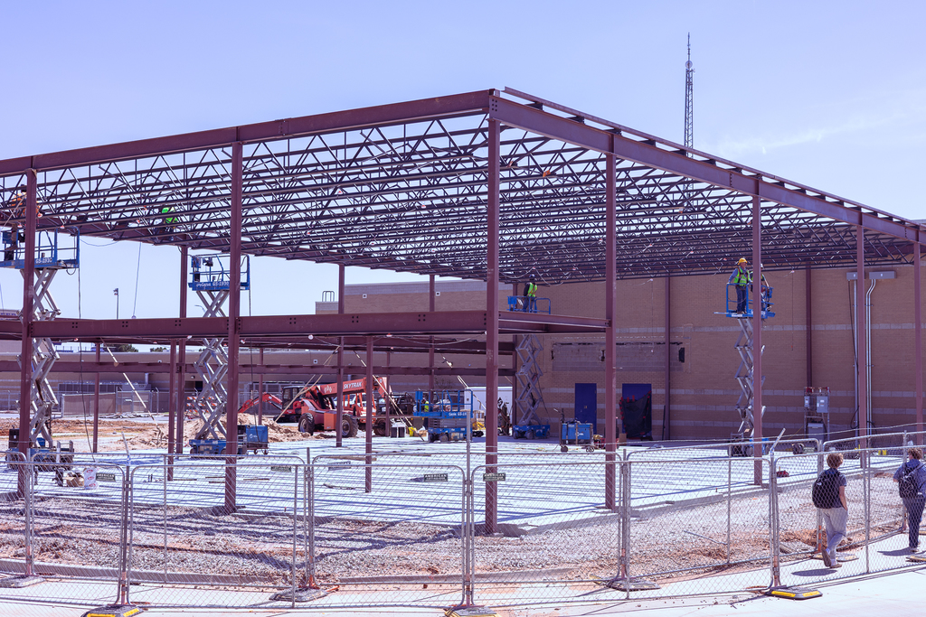 Steel framework of the Wylie High School band expansion rises from a concrete foundation as multiple workers operate lifts to install beams, with construction equipment and fencing surrounding the site.