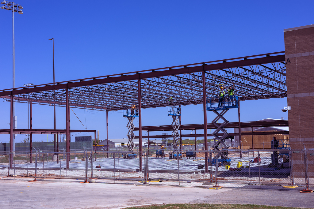 Workers on elevated lifts assemble the steel structure of the new band expansion at Wylie High School, with the building frame extending outward beside existing campus facilities under a clear blue sky.
