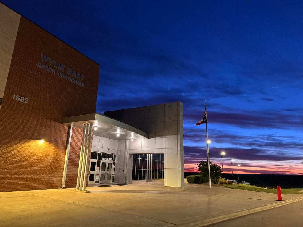 Front entrance of Wylie East Junior High School at dusk, with the building lit by exterior lights, a Texas flag on a flagpole, and a colorful sunset sky in the background.