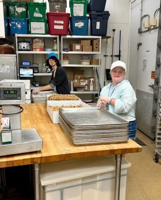 Two students working in a kitchen workspace, preparing trays of baked goods at a stainless steel table with shelves of supplies in the background.